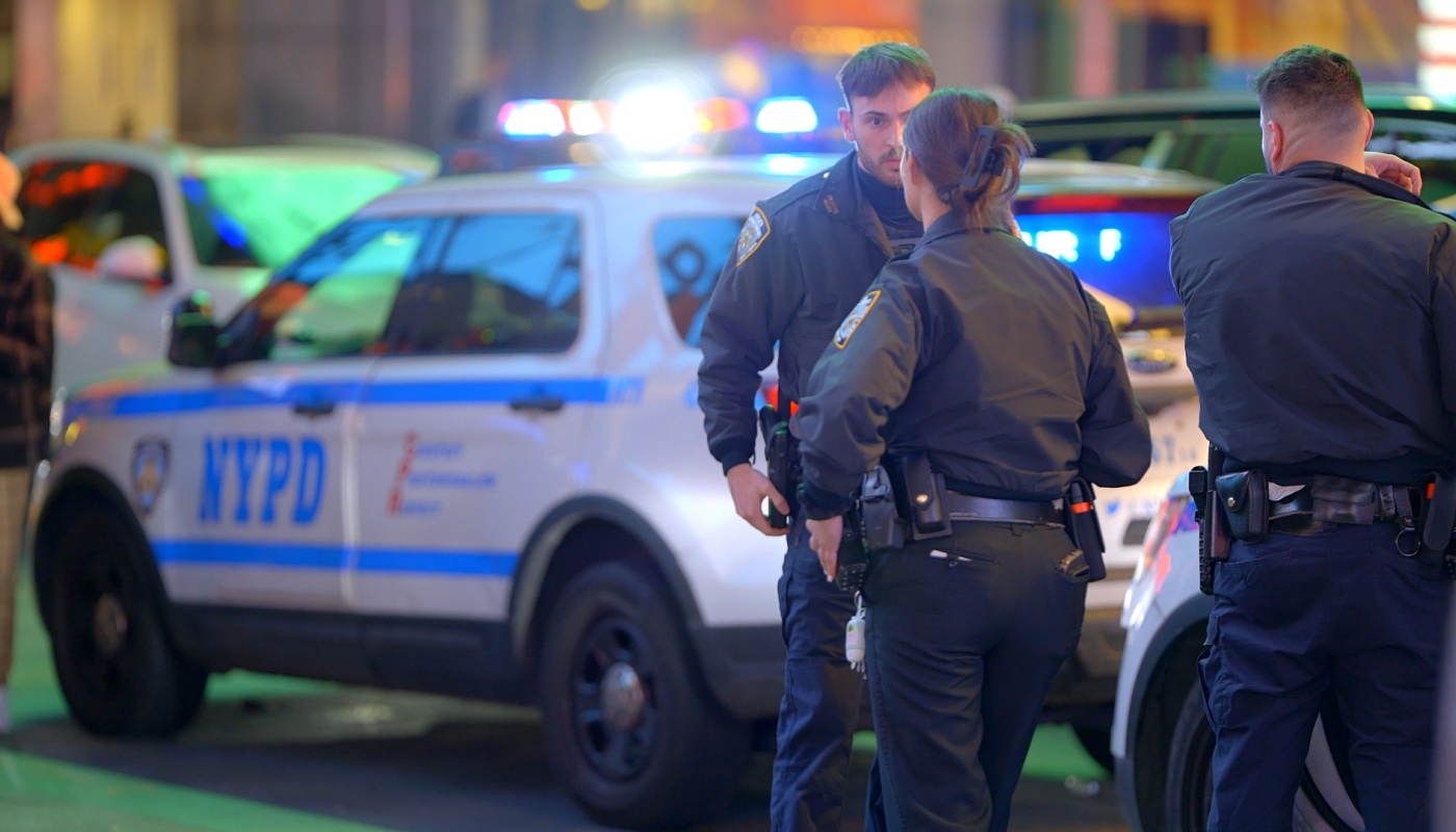A New York police car parked with the lights on, and three police officers standing next to it.
