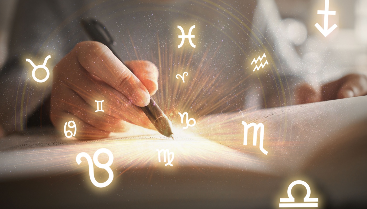 A hand holding a pen over a notebook, with Zodiac signs floating from the pages.