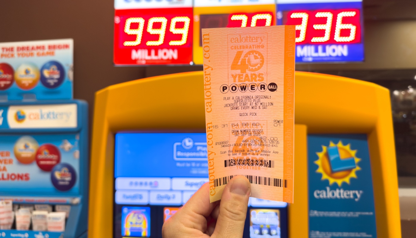 A hand holding a Powerball ticket in front of a sign displaying lottery jackpots.