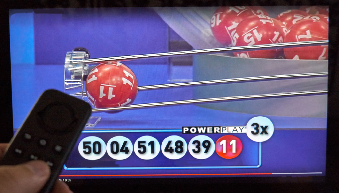 A hand holding a remote control, in front of a TV where you can see a Powerball draw being held.
